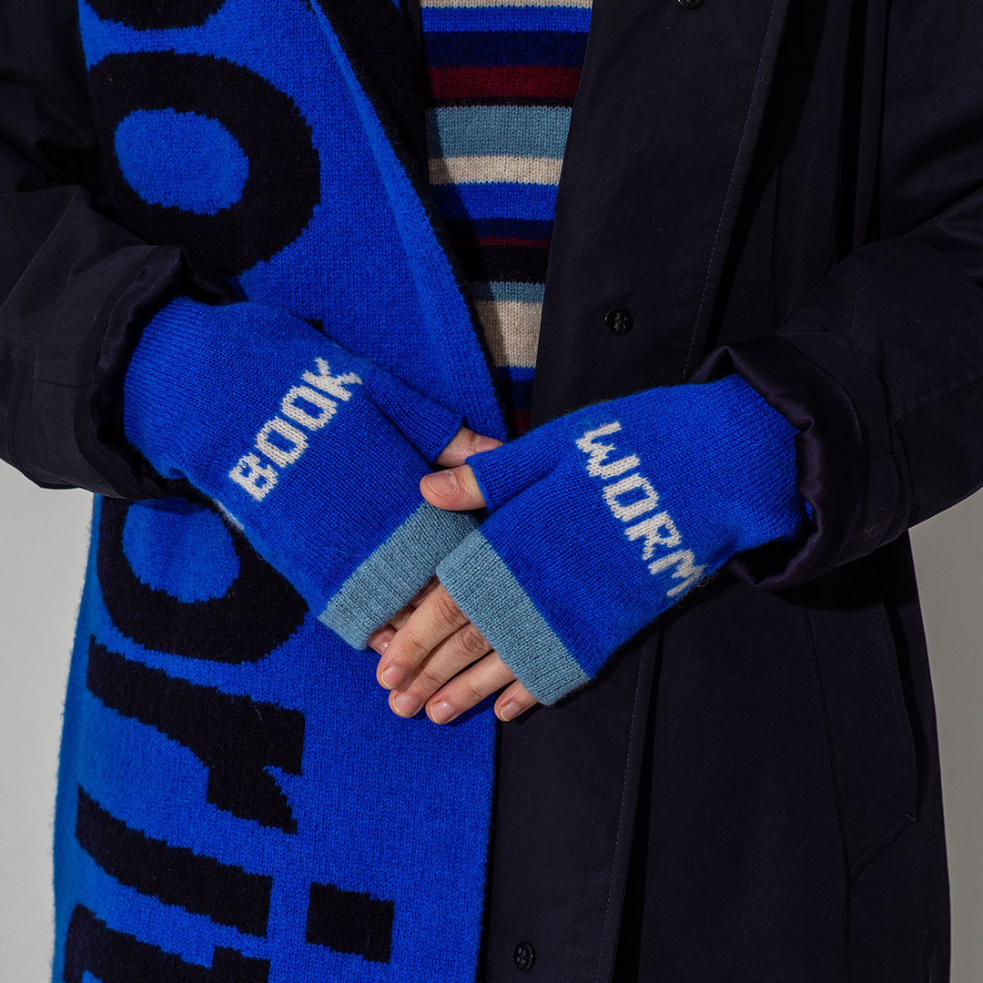 A woman in a denim shirt is holding her hands in front of her, forming a triangle with her fingers and thumbs. The hands are wearing red and pink mittens with the word 'feminist' knitted into the pattern across the knuckles.