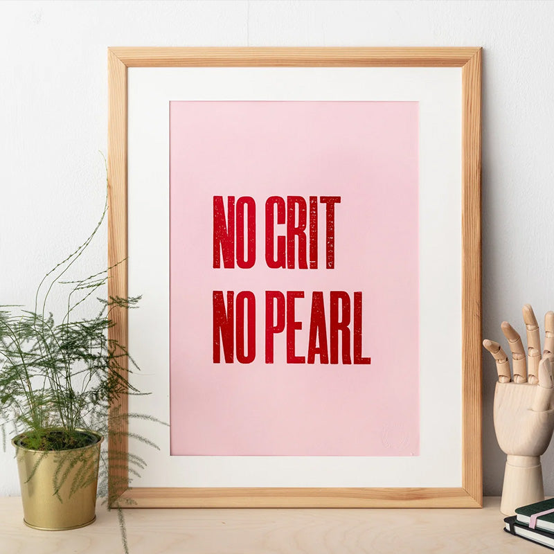 An art print with the slogan 'No Grit No Pearl' in red text on a pink background is framed. On one side is a potted plant, on the other an artist's wooden hand.