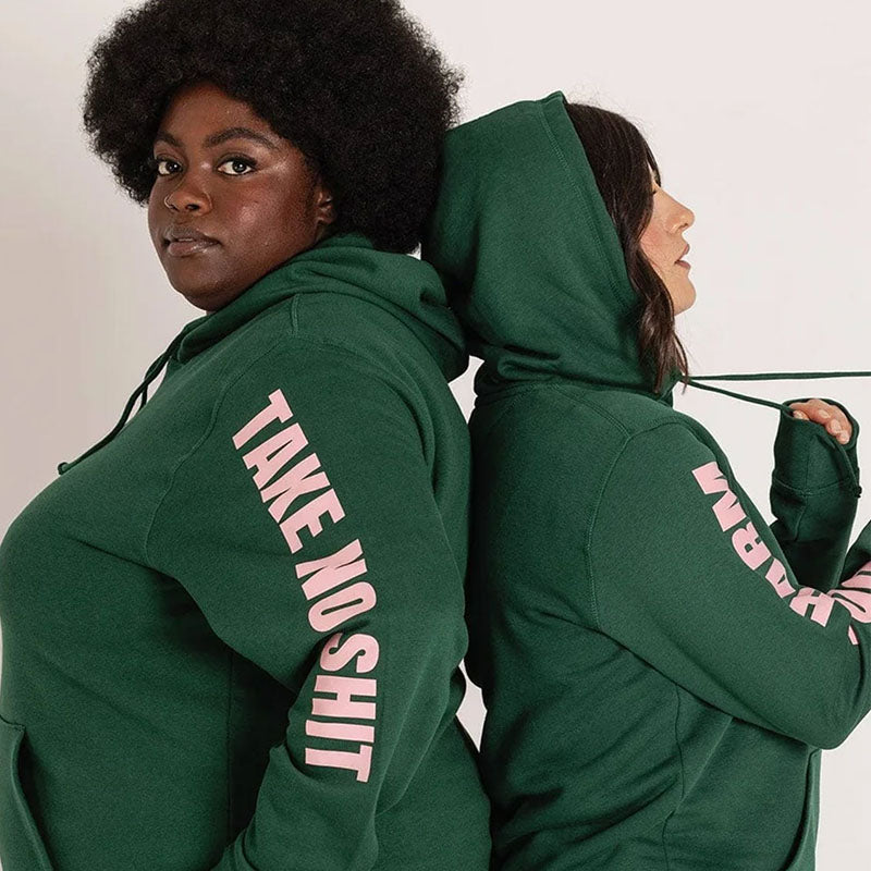 Two women stand back-to-back wearing green hoodie sweatshirts with the text 'Do No Harm Take No Shit' printed in pink along the arms.
