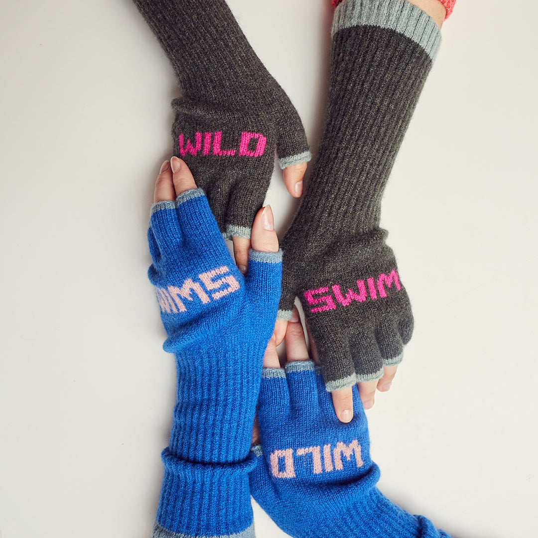 A woman in a denim shirt is holding her hands in front of her, forming a triangle with her fingers and thumbs. The hands are wearing red and pink mittens with the word 'feminist' knitted into the pattern across the knuckles.