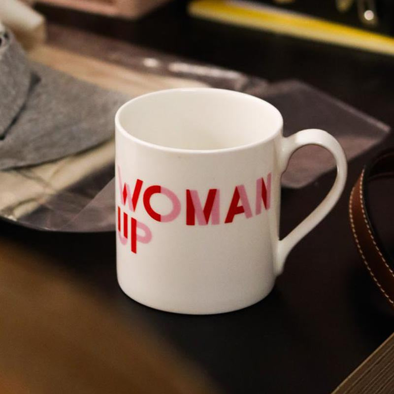 A white mug, with the text 'Woman Up' printed across it, sits on a table..
