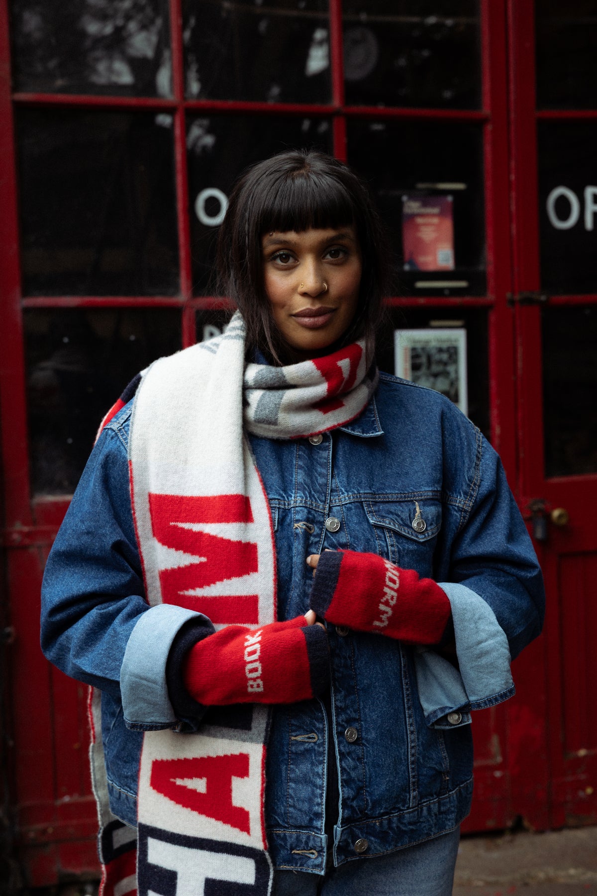 A woman wearing a blue denim jacket, a white and red striped scarf, and red knit gloves with 'HAMM' printed on them, standing in front of a red telephone booth.