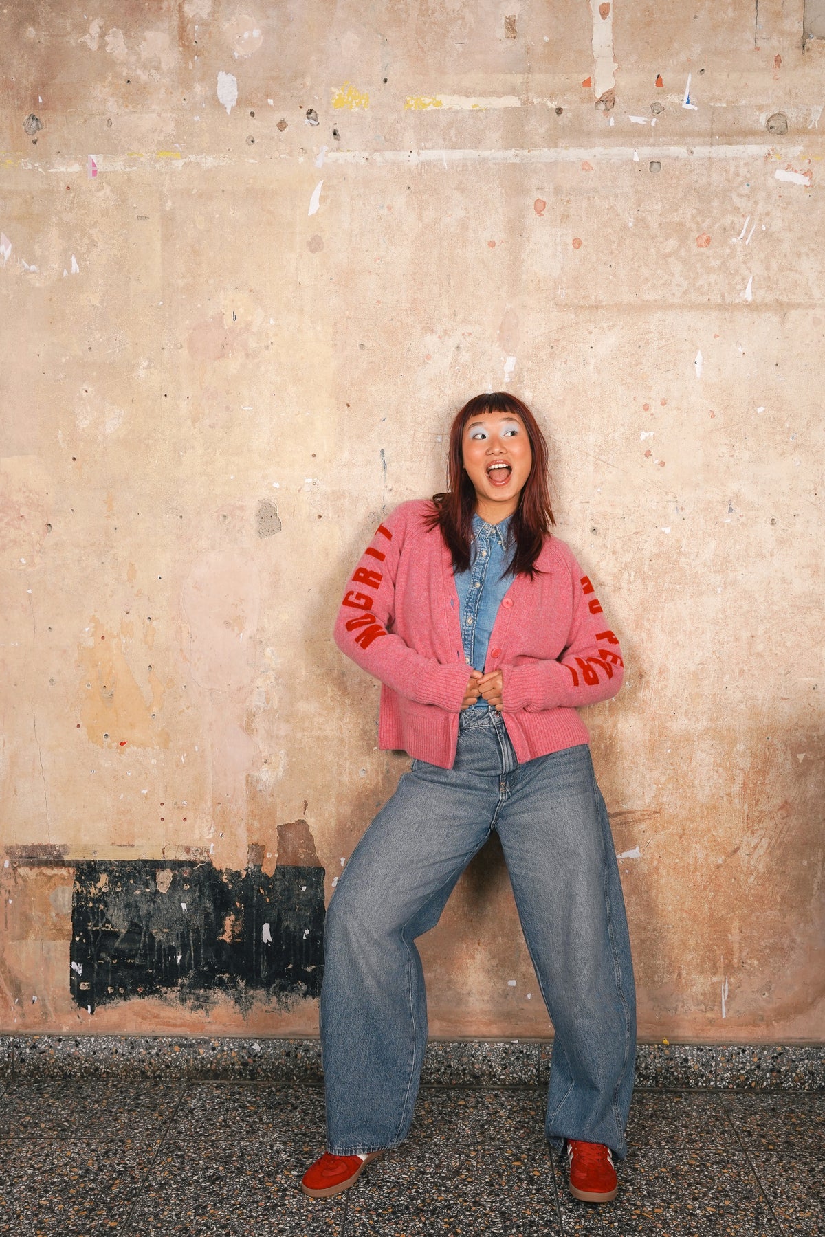 A woman posing in front of a textured concrete wall, wearing a pink cropped jacket with red lettering on the sleeves, a blue denim shirt, wide-leg blue jeans, and red shoes.