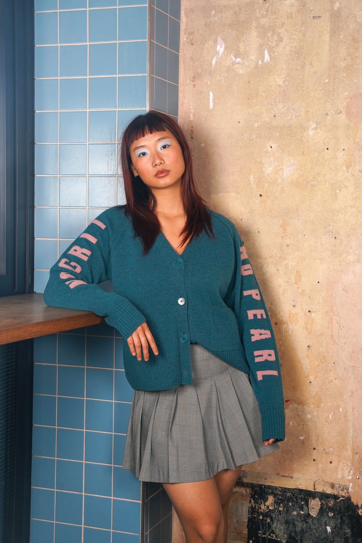 A woman wearing a teal button-up cardigan with pink 'PEARL' lettering down the sleeves, paired with a gray pleated mini skirt, leaning against a textured wall next to blue tiled panels.