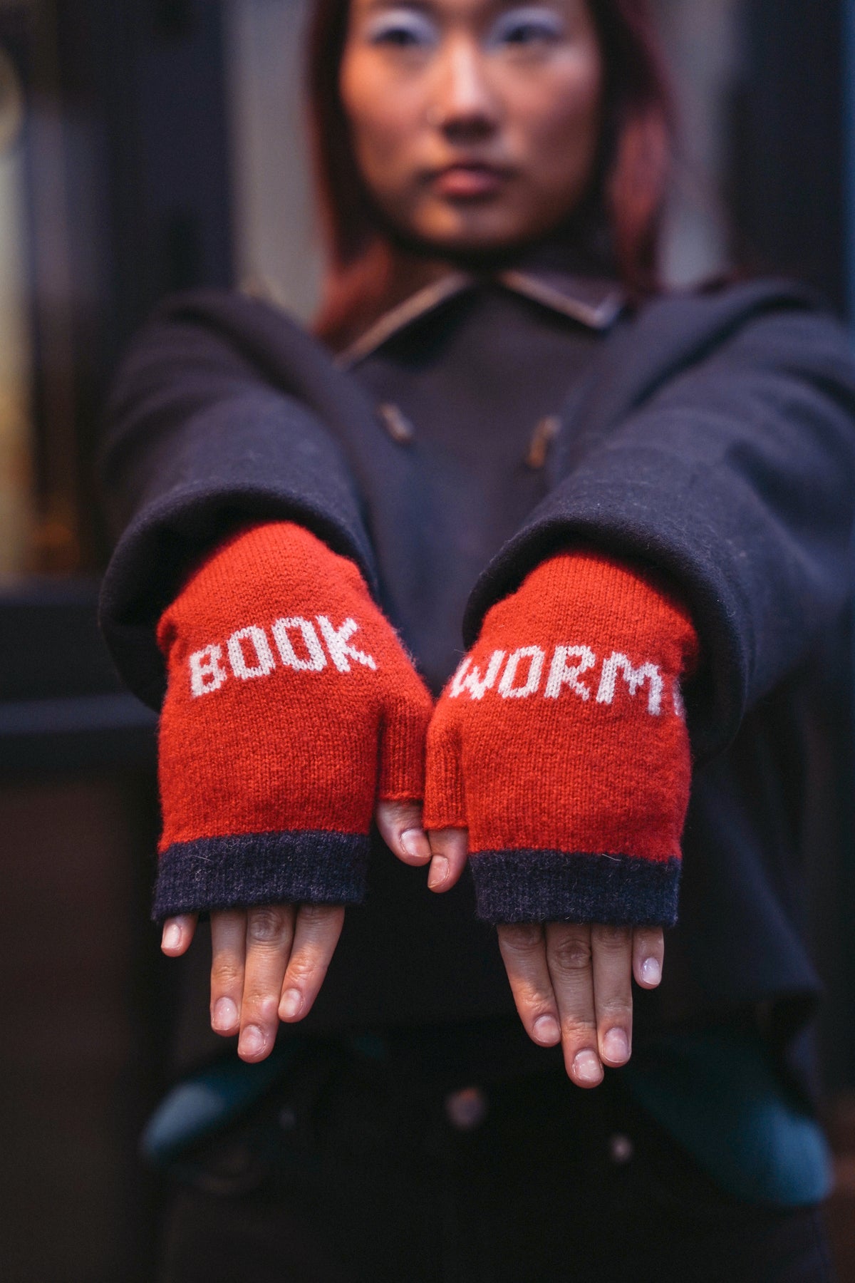 A person wearing fingerless knit gloves with the words 'BOOK WORM' in white lettering on bright red palms, shown against a dark blurred background.